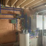 Mechanical room with blue and green manifold piping, boiler units, control panel, and white heating tanks