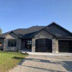 Modern stone home with dark roof, black garage doors, and mature landscaping