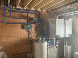 Mechanical room with blue and green manifold piping, boiler units, control panel, and white heating tanks
